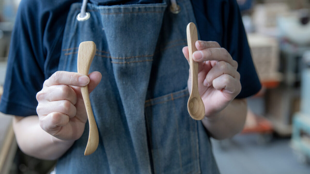 木製ジャムスプーン『saji』と『nairo』を手に持つ女性の木工職人。背景には木工工房の作業スペースが広がっている。