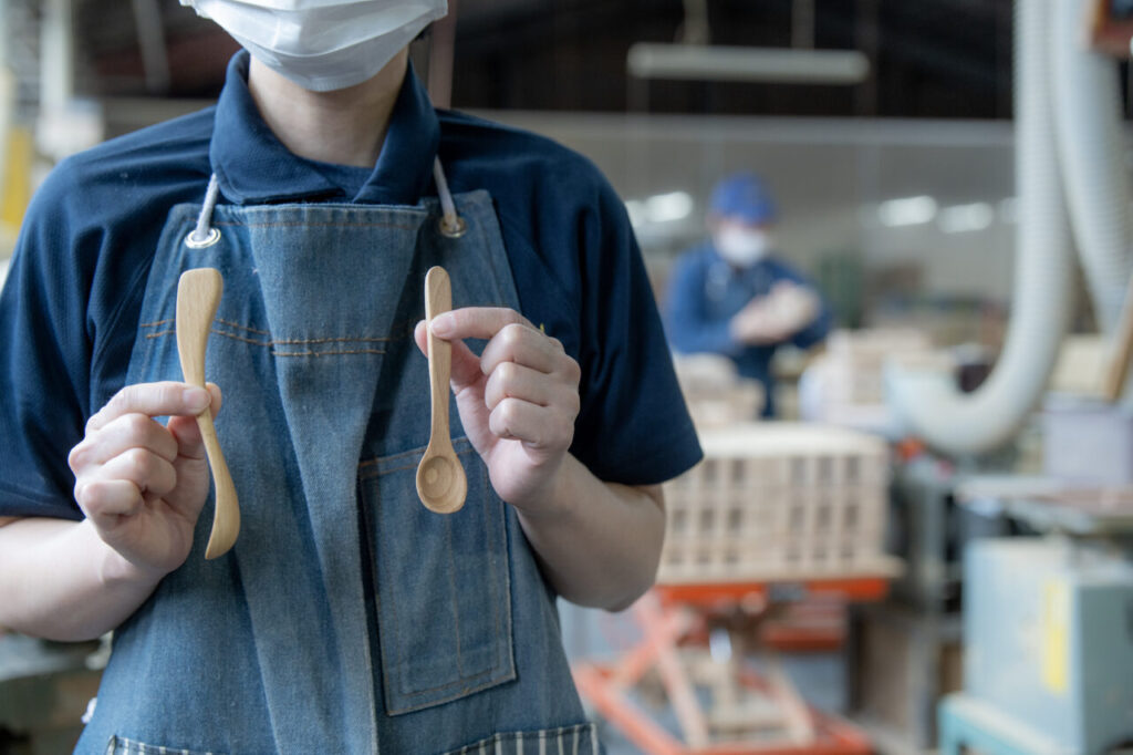 木製ジャムスプーン『saji』と『nairo』を手に持ち、集中して作業する女性の木工職人。背景には木工工房の作業台や道具が整然と並んだスペースが広がっている。